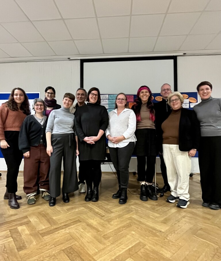 Eine Gruppe von zwölf Personen steht in einem Innenraum vor einer Leinwand und mehreren farbigen Moderationskarten, die an Pinnwänden befestigt sind. Alle schauen in die Kamera und lächeln. Die Gruppe besteht aus Menschen unterschiedlichen Alters und unterschiedlicher Erscheinung; einige tragen Brillen, eine Person hat pink gefärbte Haare. Sie stehen in einem Halbkreis auf hellem Holzfußboden. Die Atmosphäre wirkt warm, kollegial und konzentriert.
